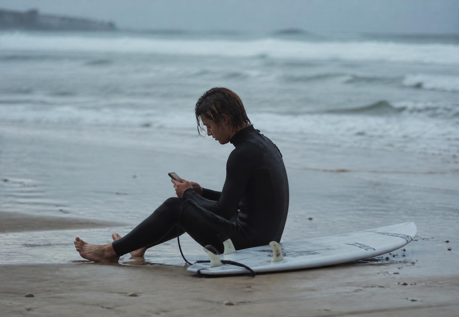 A surfer sitting on the sand checking their phone after a session, board beside them